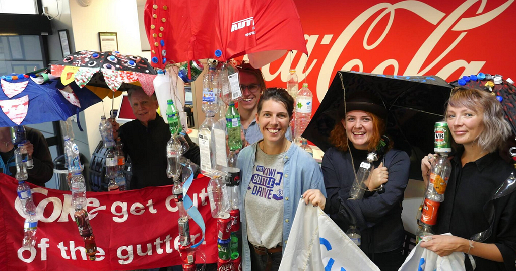 Holly and volunteers holding plastic bottle creations and signs. A banner reads ‘Coke get out of the gutter’