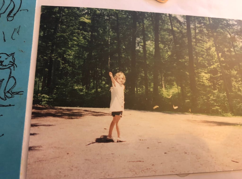 A young white female child looks into the camera. She is outside in a parking lot and there are trees behind her. In the foreground with her there are butterflies. She is extending her arm towards the person taking the picture.