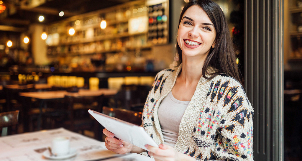 A smiling woman is working on her iPad in a cafe. Photo by Ivan Pantic on Canva.