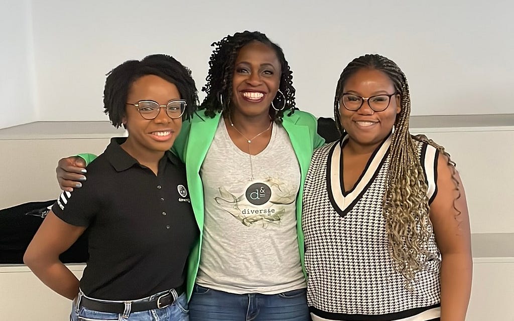 Three black women, with their arms around each other, stand side by side and are smiling to camera.