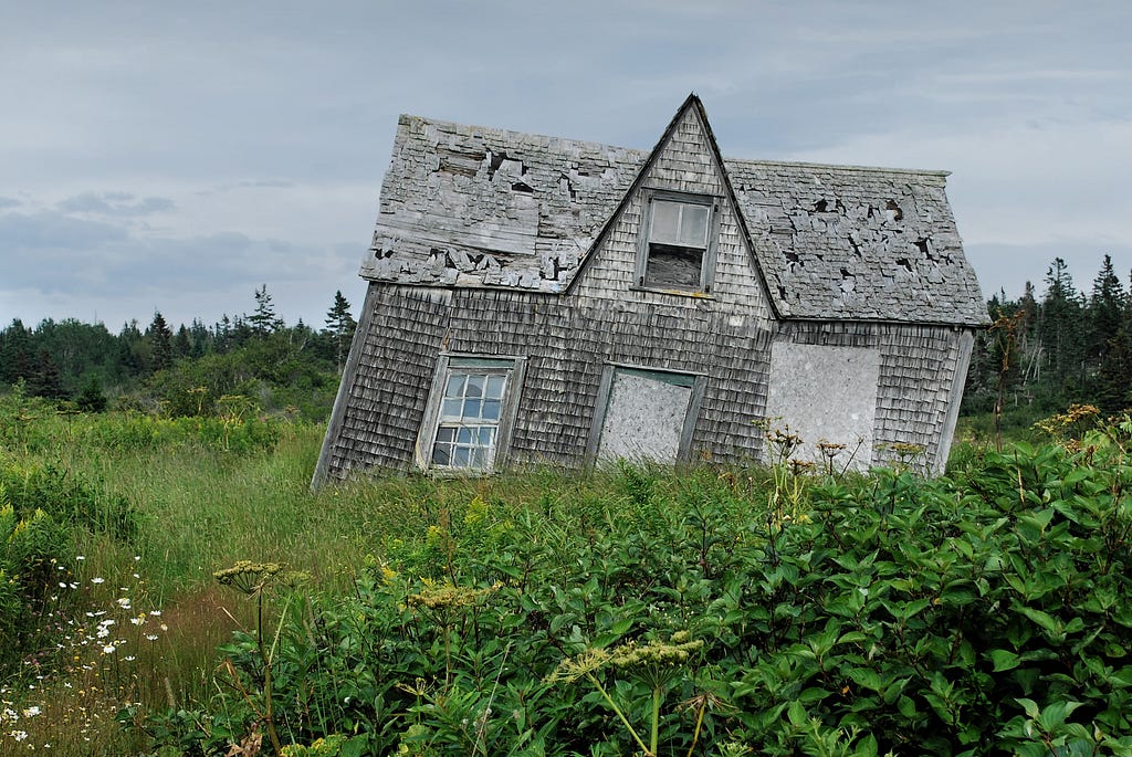 An old run down gray wooden house that leans entirely to the right with holes in the roof and boarded up doors