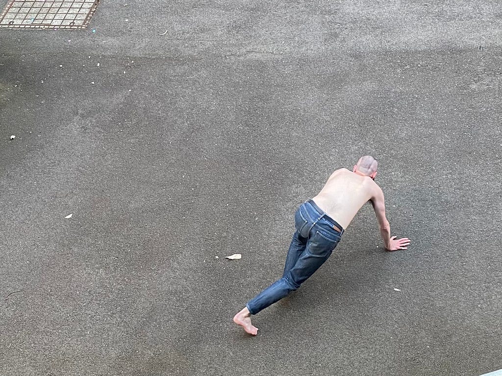 A person in blue-grey jeans walks around on all fours like a dog. They are shirtless, and the ground beneath them is concrete. They have their back to us and we cannot see their face.