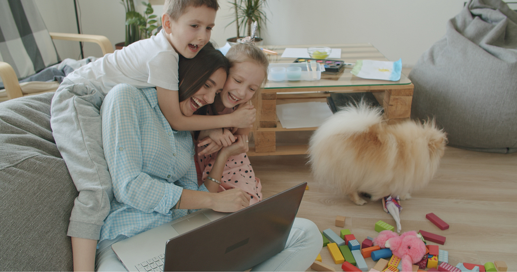 a laughing mother working on her laptop surrounded by her children hugging her in their play room.