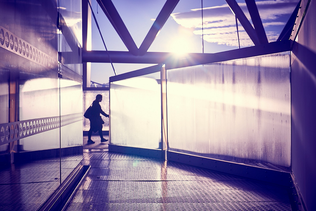 Image: the silhouette of a woman with a big handbag passing between opaque sheets of glass on a pedestrian bridge in Oslo