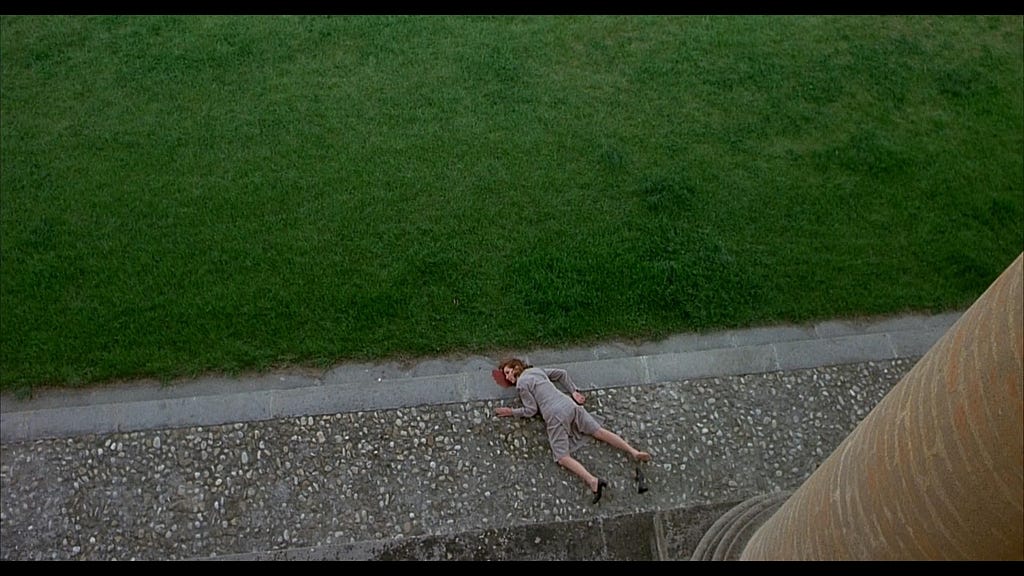 A shot from Salo which shows a woman, the Pianist, crumpled on the ground with blood coming out of her head, clearly dead. The very edge of the window she threw herself out of can be seen at the bottom of the shot, and the top two-thirds is taken up with green lawn. Her body lies between the lawn and the window-ledge.