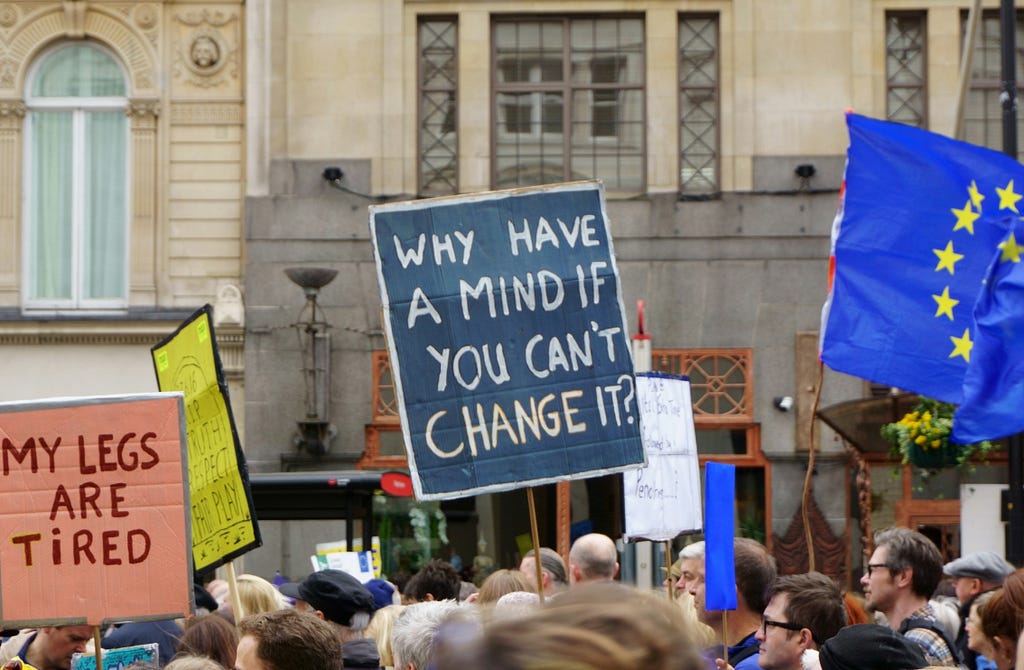 Photo of a protest. There people holding up signs which read (from left to right): “My legs are tired” and “Why have a mind if you can’t change it?”