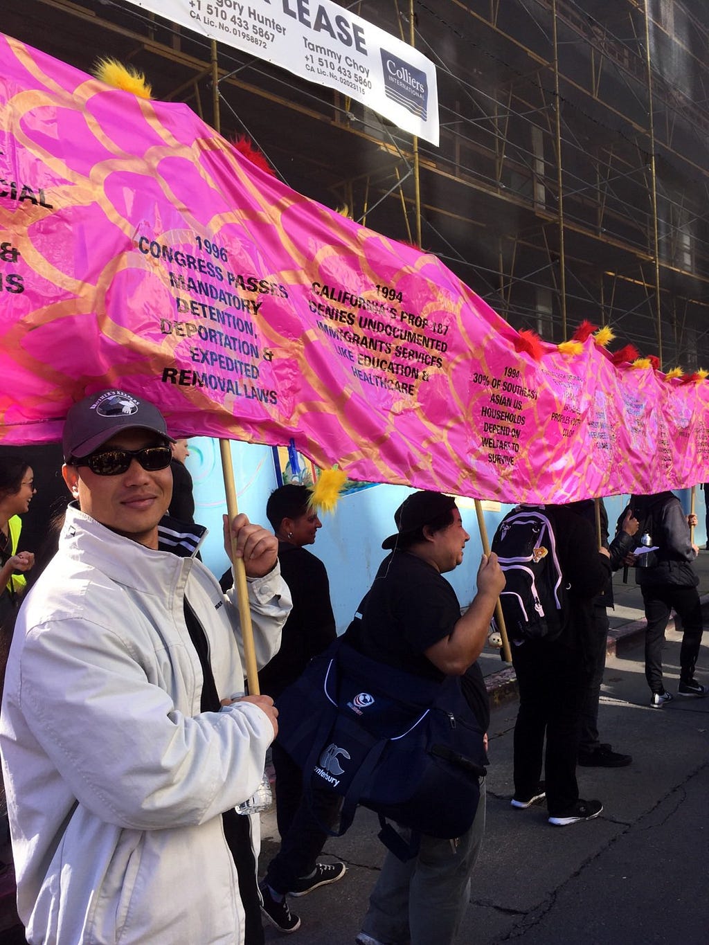 A group of Asian people of different ethnicities carry a colorful pink fabric banner on poles, with dates & historic blurbs.