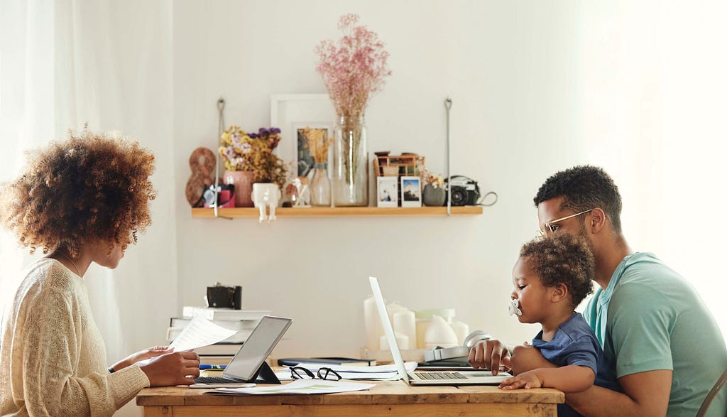 Young couple working from home sitting in the kitchen — woman using a digital tablet and man using a laptop with their toddle