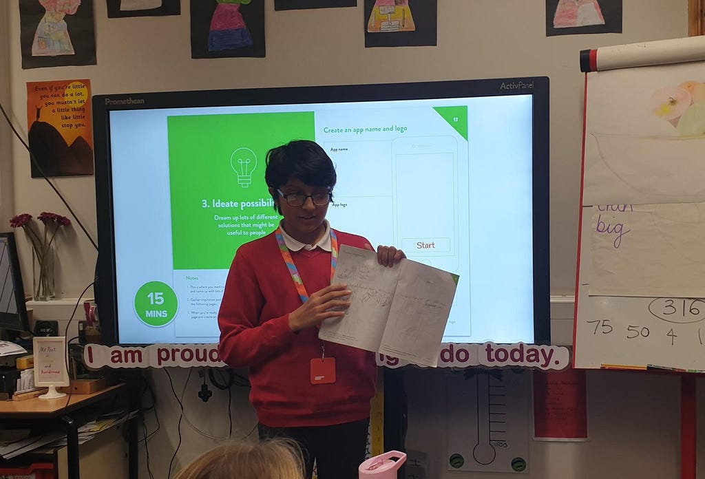 A Year 6 wearing a school uniform and Design Club lanyard holding an app design workbook in front of a screen, at the front of the classroom showing the ‘Ideate possibilities’ page from the workbook.