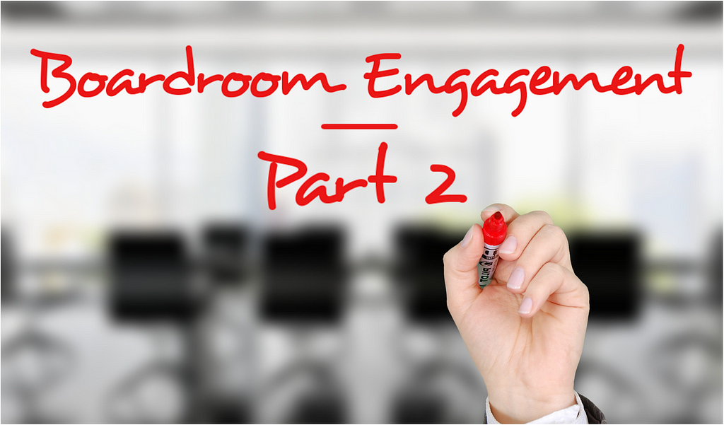 A hand holding a red pen writing on the screen the words “boardroom engagement — part 2” with a blurred picture of a boardroom in the background.