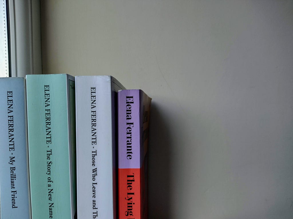 Four novels on a windowsill. They have blue, green, white, and a red and purple spine. They are all written by Elena Ferrante.