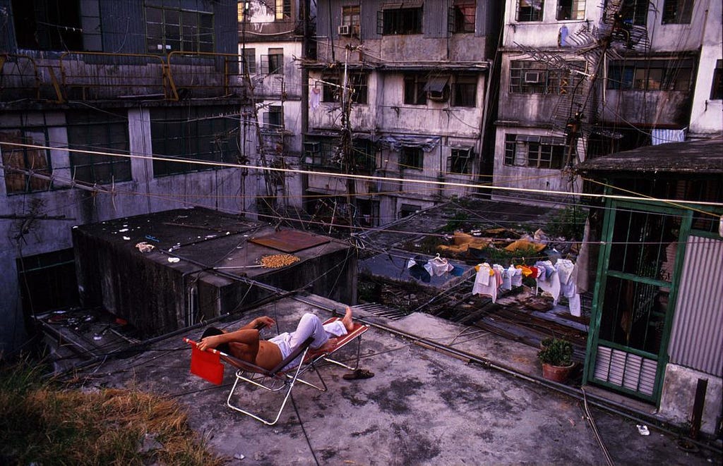 A shirtless man reclines in a folding deck chair on a rooftop in Kowloon Walled City, Hong Kong. Around him, a dense web of electrical cables stretches between the surrounding dilapidated apartment buildings. A clothesline with laundry hangs nearby, and a small potted plant sits by a green-framed window. The rooftop is littered with debris, and grain is spread out to dry on a nearby surface.