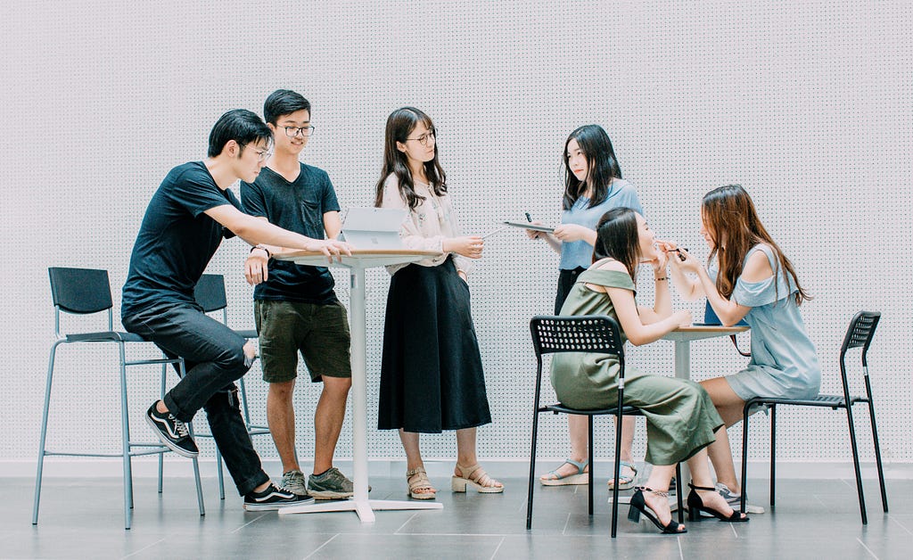 Six young adults sitting/standing at tables and interacting.