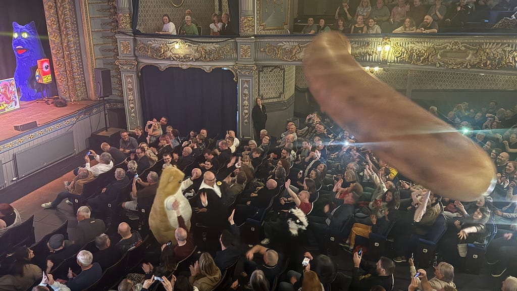 A large inflatable sausage is shown above the audience of the Harry Hill show, with Harry and company standing in the aisle