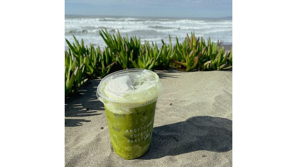 A matcha drink sits in the sand with the ocean waves and sea grass behind it