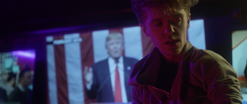 A man with blonde hair and wearing a gray hoodie faces right. A tv in the background shows Trump at a podium.