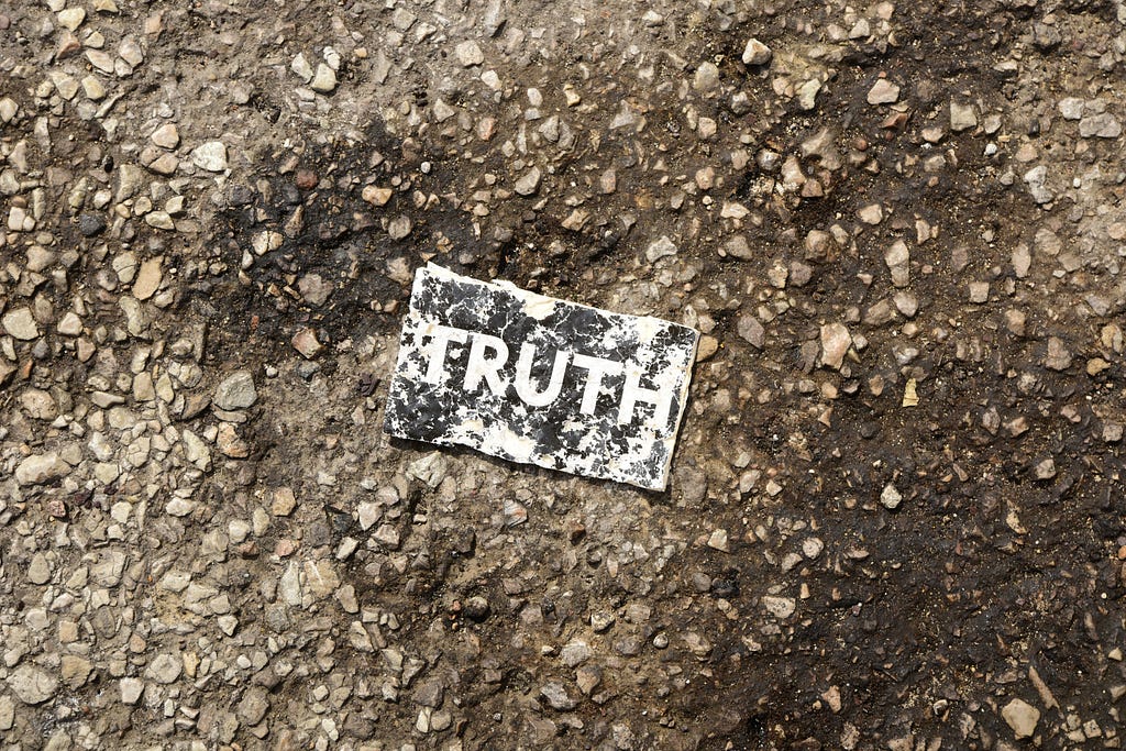 A black tattered piece of paper with the word TRUTH in white text on asphalt