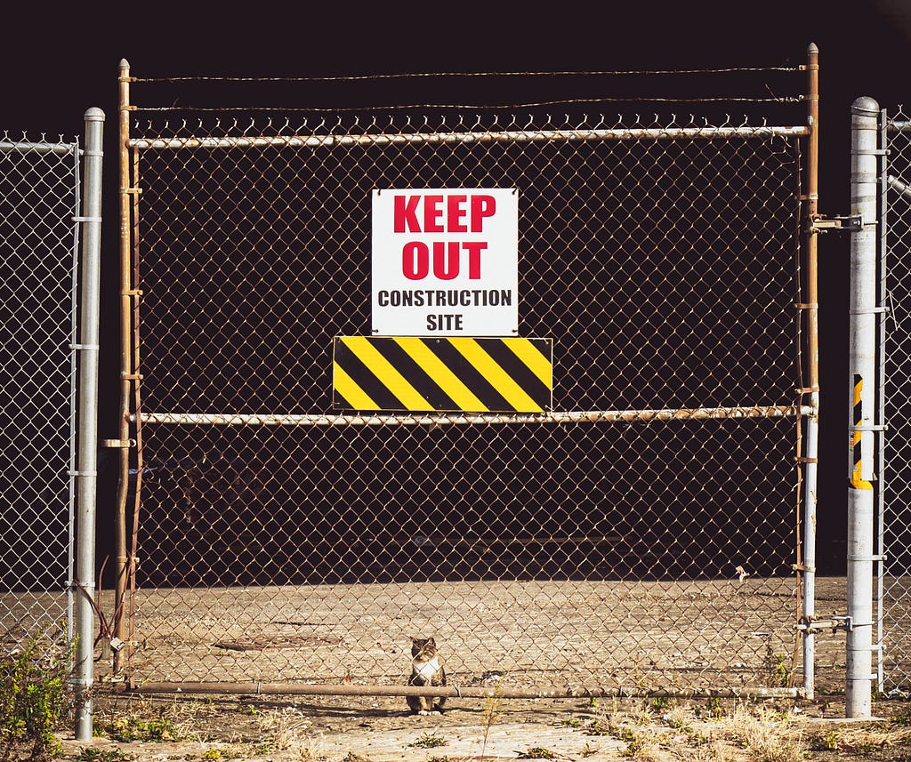 A cat sits behind a fence, peering through a gate with a sign that warns “keep out”