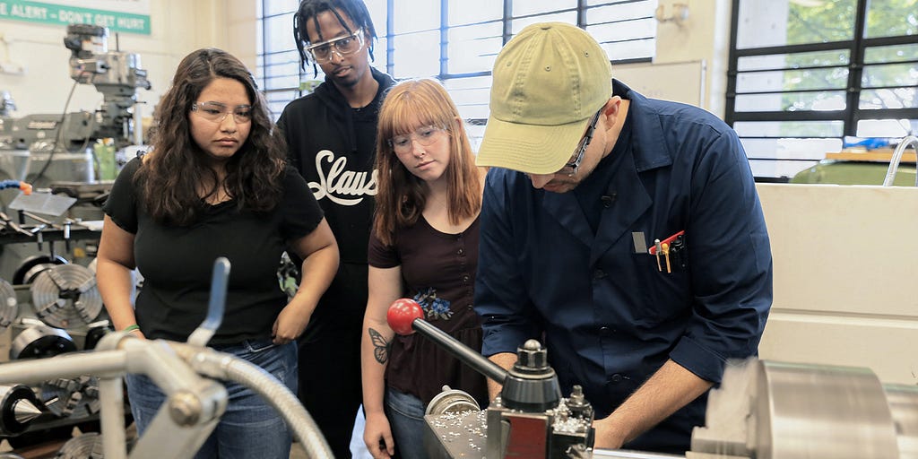 Three students watch as an instructor operates a lathe.