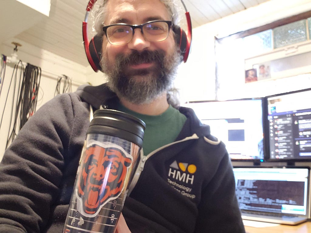 A man holding a drinks flask in front of his work station, wearing an HMH hoodie