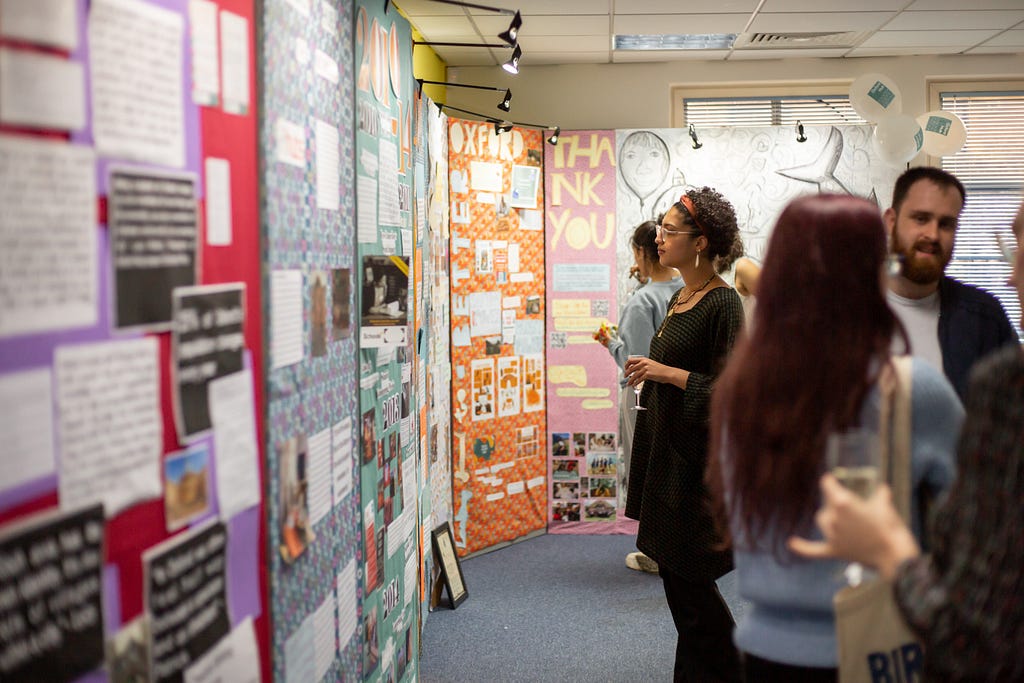 A woman looking at a brightly-coloured exhibition. People mill around, chatting and looking at the exhibition around her.