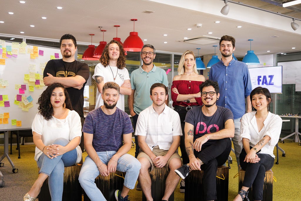 Grupo de pessoas em primeiro plano, sendo 5 em pé e 5 sentadas em frente em uma sala colorida.