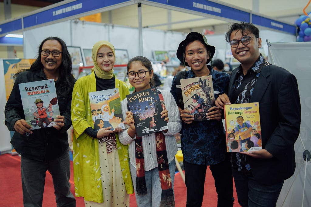 Five adults in a row, holding their books. On the left is a man with long black hair and glasses. Second to the left is a woman in a bright yellow hijab and matching sweater. In the middle is a younger woman with glasses, a long gray shirt, and a shim scarf. Second to the right is a younger man in a hat and dark-blue button-down. On the far right is a man with a wisp of hair falling on his forehead wearing glasses and a suit. All five are South-Asian.