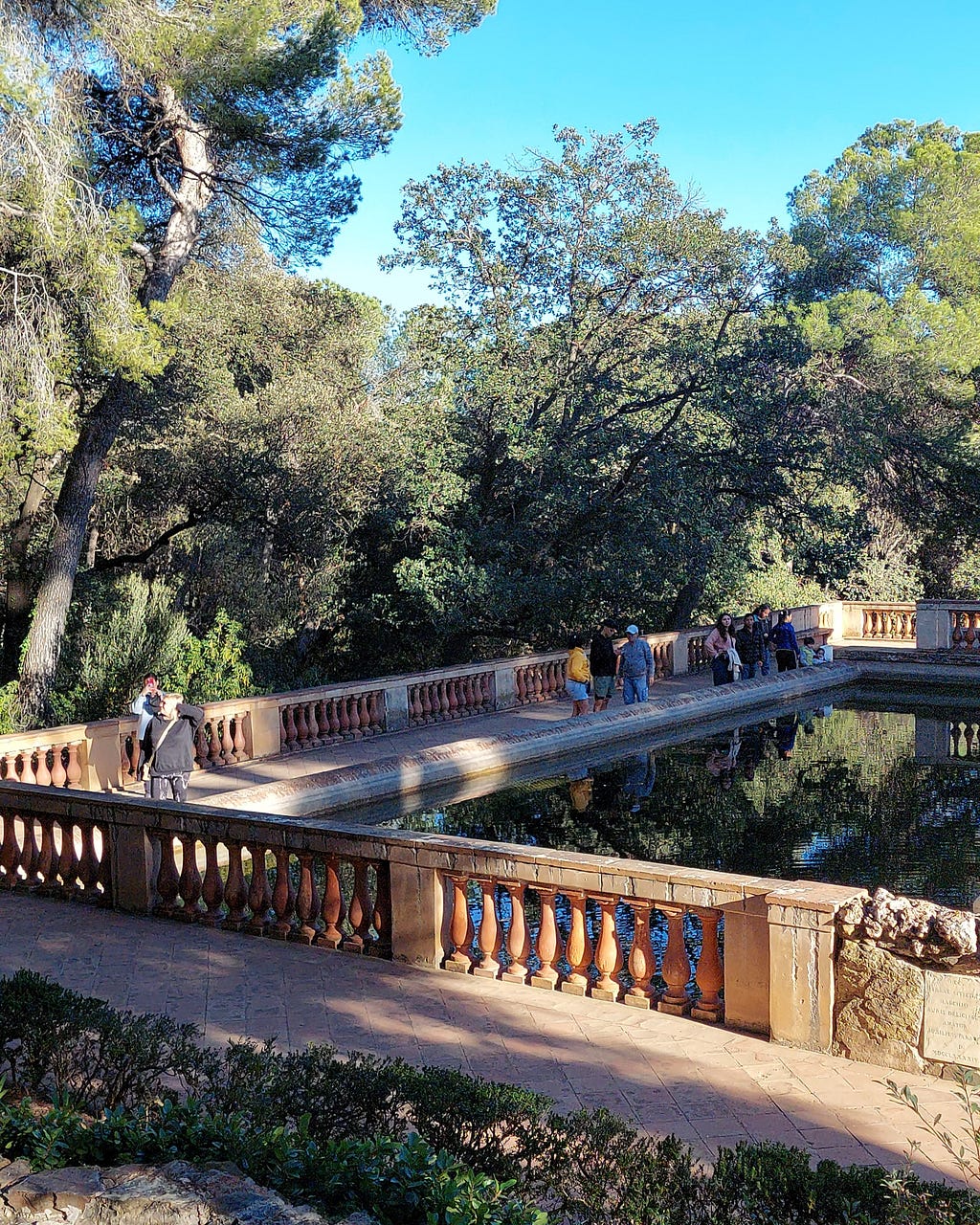 View of the historic pavilion of the Horta Labyrinth Park, with an artificial lake at the back. In the image, we see some tourists, many trees and the sky.