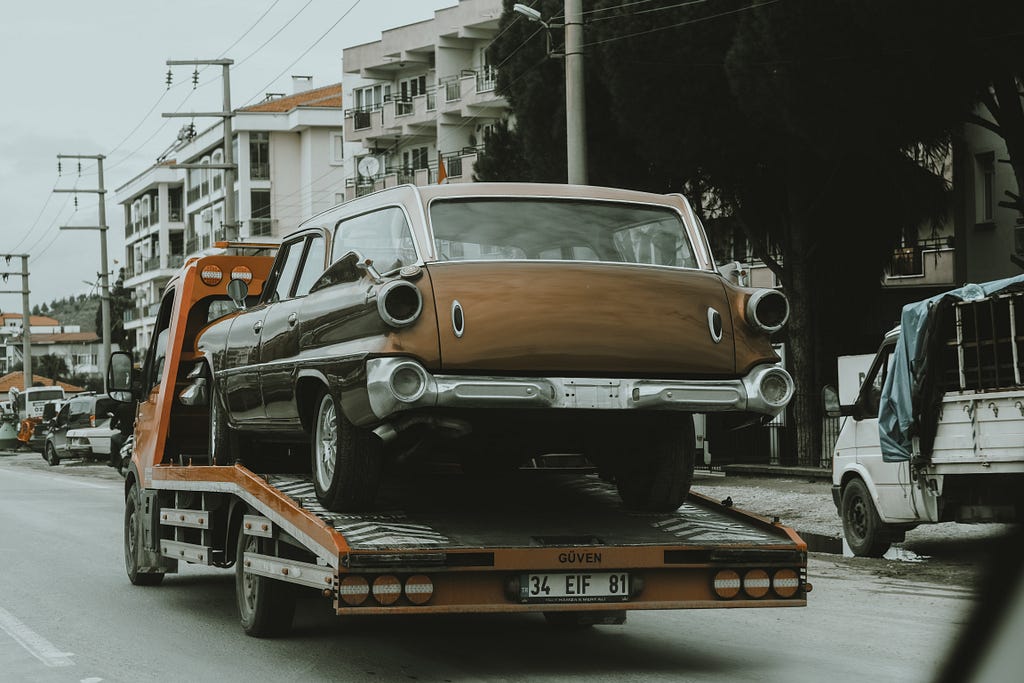A old brown sedan is on the back of a red tow truck being towed away on a city street in Europe.