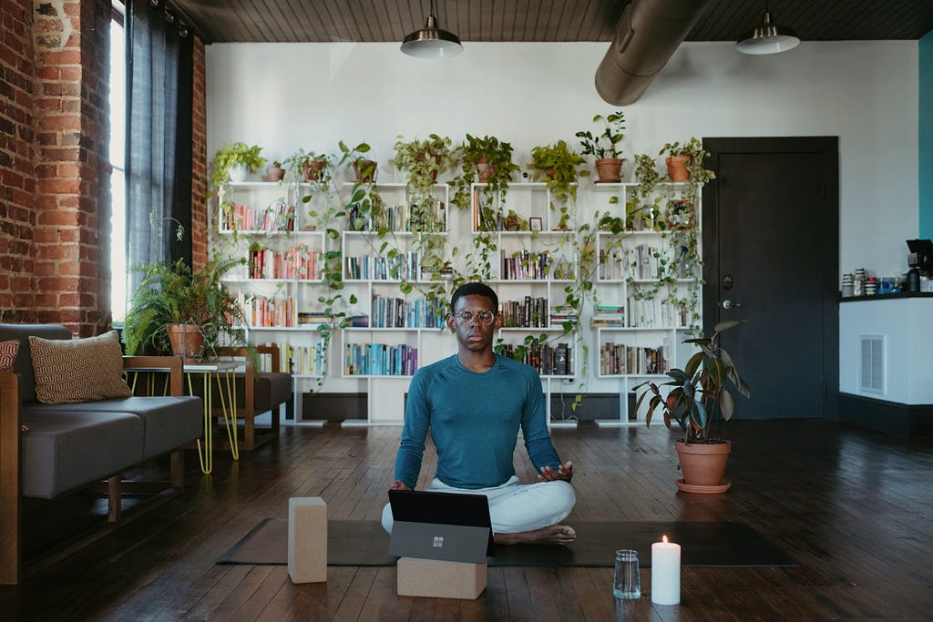 Man meditating in a room filled with books and plants and a burning candle.
