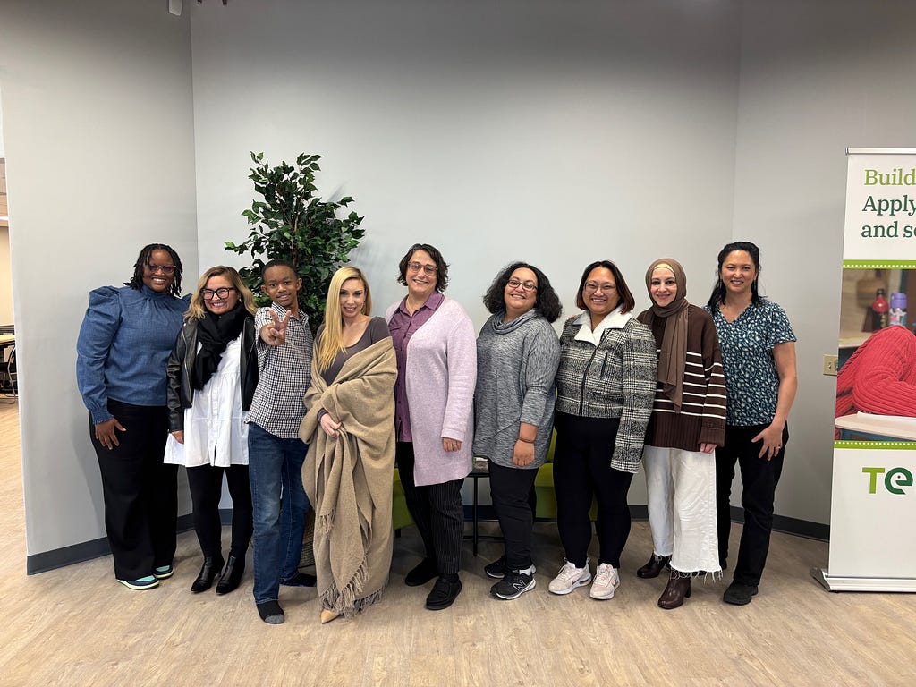 Dr. Lisette Torres-Gerald with seven other people. They are inside a building standing in front of a talk green plant. Everyone is smiling and one person is holding out the peace sign.