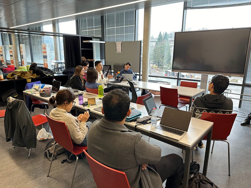 People sit in red chairs in front of a few tables, with laptops, water bottles, and other general workshop materials; they sit inside a room facing large glass windows, with green trees and overcast gray skies in the background.