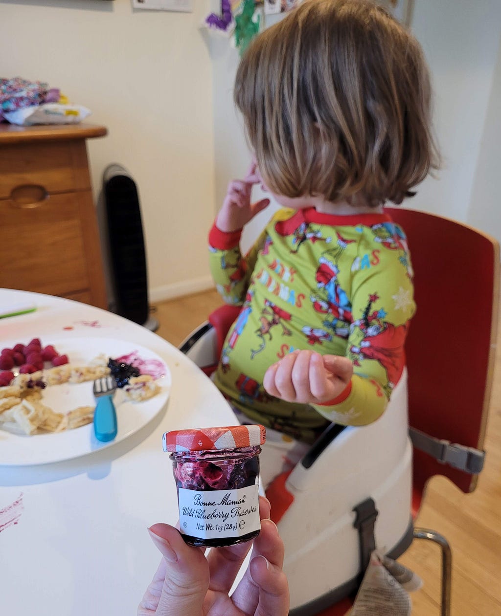 A small child wearing green and red pajamas sits at the table, facing away from the viewer. She has a plate of unfinished waffles and raspberries in front of her. The author holds a tiny jar of jam in the foreground.