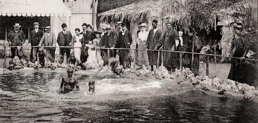 Circa 1904. White patrons watch from behind a guardrail as people of African descent wade in a pond at a “human zoo”