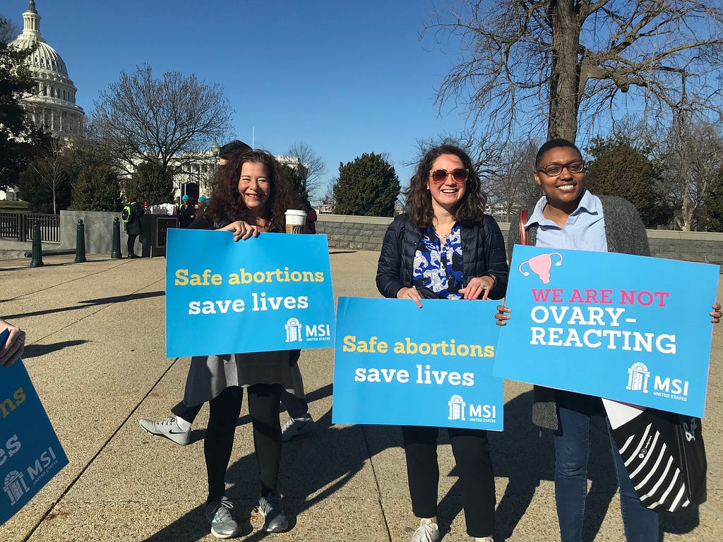 Three women holding signs, two of which say “Safe abortions save lives,” the other reads, “We are not ovary-acting”.