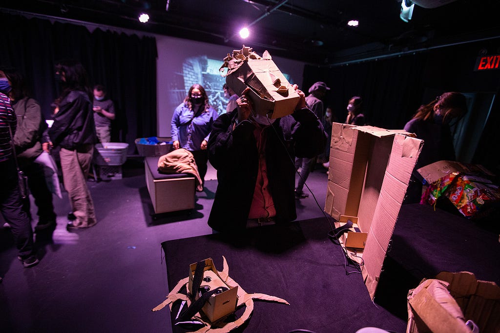 A visitor tries on a square-ish cardboard mask from a table of differently shaped masks, while others in the dimly lit room look at projected scenes of different peoples’ “memory enclaves” from the week of workshops.