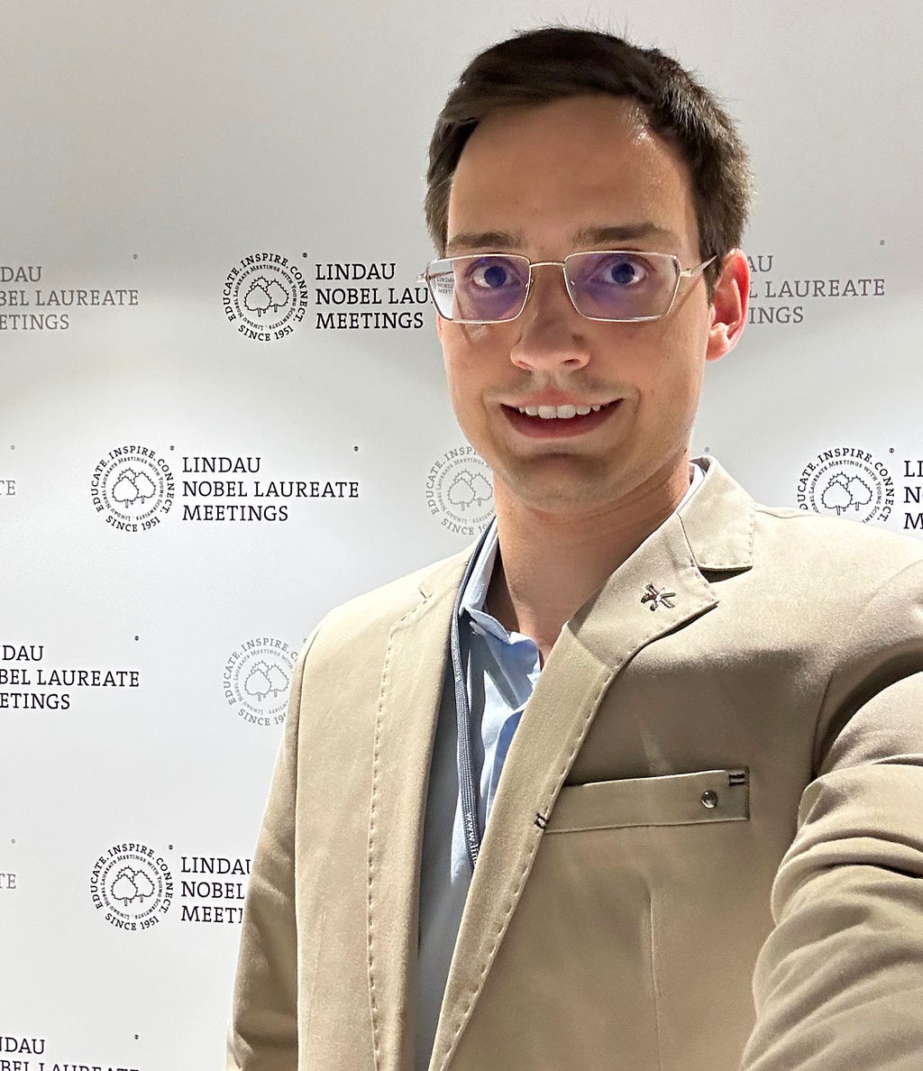 Selfie de Gustavo Rosa Gameiro, ele é um homem branco jovem, usa óculos, paletó beje e camisa azul e ao fundo está uma parede com o marca e texto do “Lindau Nobel Laureate Meetings”