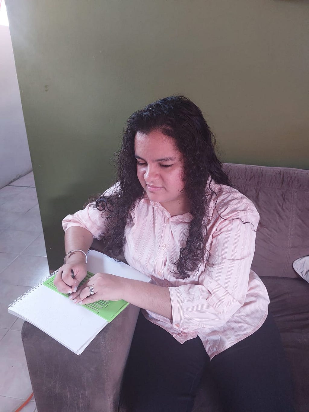 Una foto de Estefanía sentada en un sofá, escribiendo notas en un cuaderno. Lleva una camisa de color rosa claro, pantalones negros, y el cabello suelto sobre los hombros.