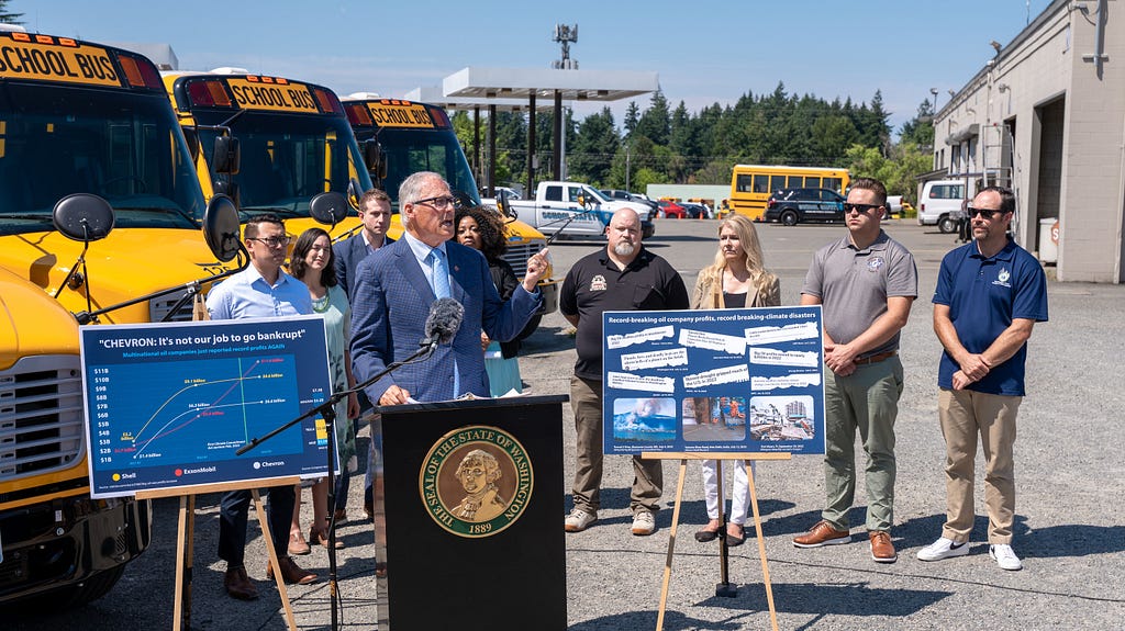 Photo of Gov. Inslee standing outdoors at a podium with a small group of people behind him. Behind the group are new electric school buses, and next to the podium are two posters — one showing increasing oil company profits and another showing a collage of headlines about oil company profits and climate-related weather events and disasters.