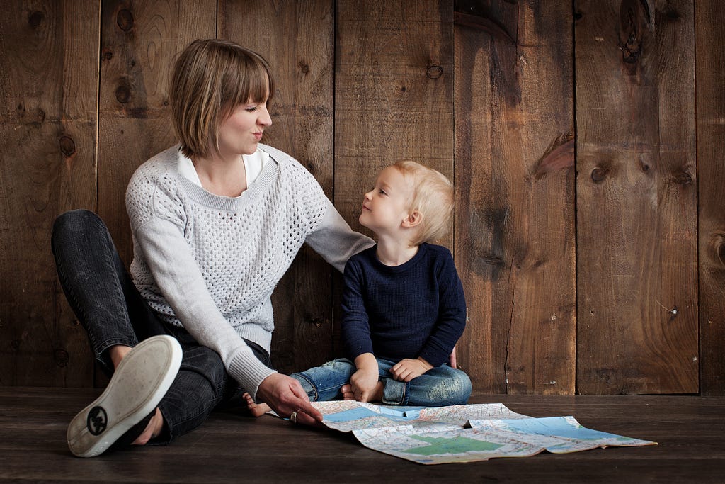 Toddler boy looks at his mother making a funny face. There is a map in front of the boy on the floor, where they both sit.