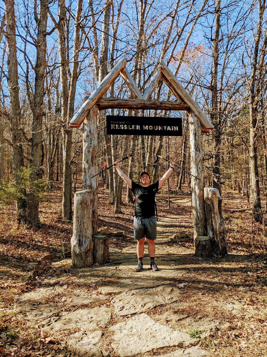 A man holding trekking poles stands under a sign that reads, “Welcome to Kessler Mountain”.