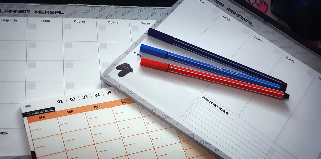 Foto com 3 blocos de papel para planejamento mensal e semanal e três canetas, uma azul, uma vermelha e uma preta.