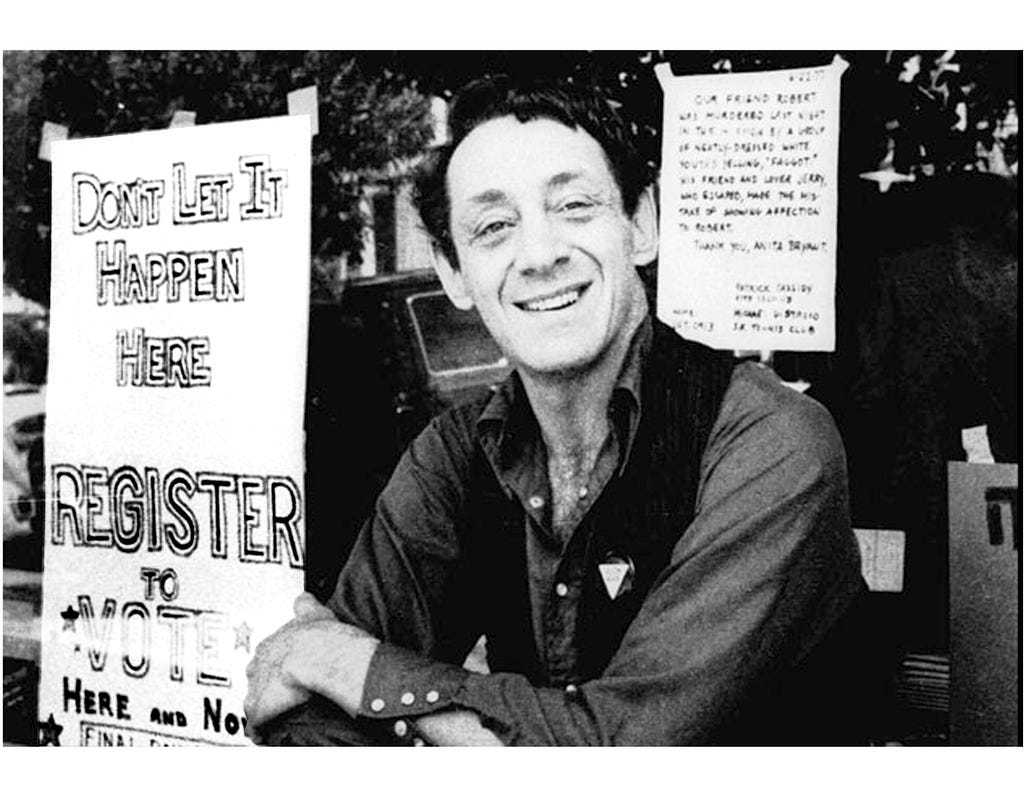 Black and white news-clipping photo of Harvey Milk, a white man with dark hair in his forties, sitting in front of gay rights signs encouraging people to vote. He is smiling into the camera.