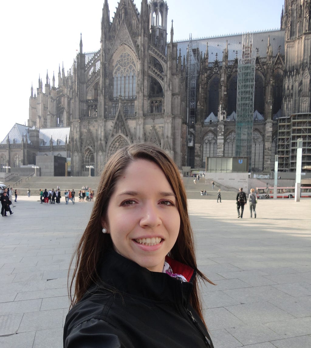 The author stands in a plaza in front of the gothic Cologne Cathedral, some of which is obscured by scaffolding.