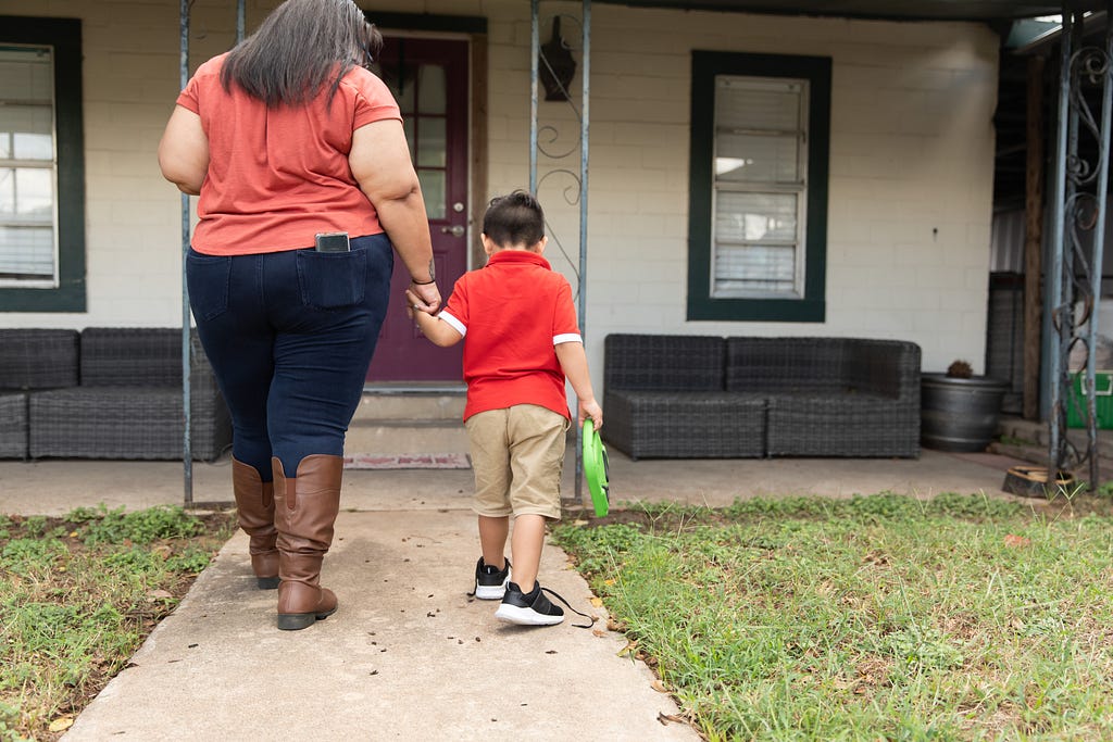 A mom holds hands with her son as they walk in their front yard into their home.