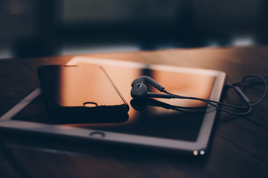 A pair of headphones rests on top of a tablet next to a smartphone on a desk. The light of the sunset shines on them through a window