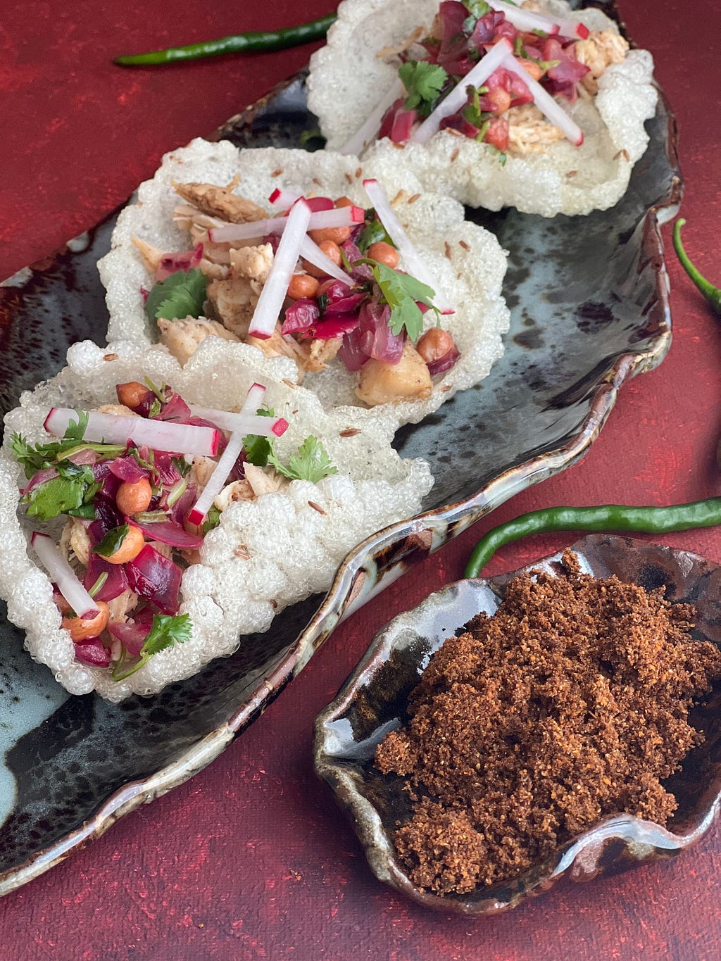Grey ‘leaf plate’ with white cracker, loaded with colorful magenta and white fillings. Small plate on side loaded with a brown powdered masala. Green chili in background. All assembled on a maroon background.