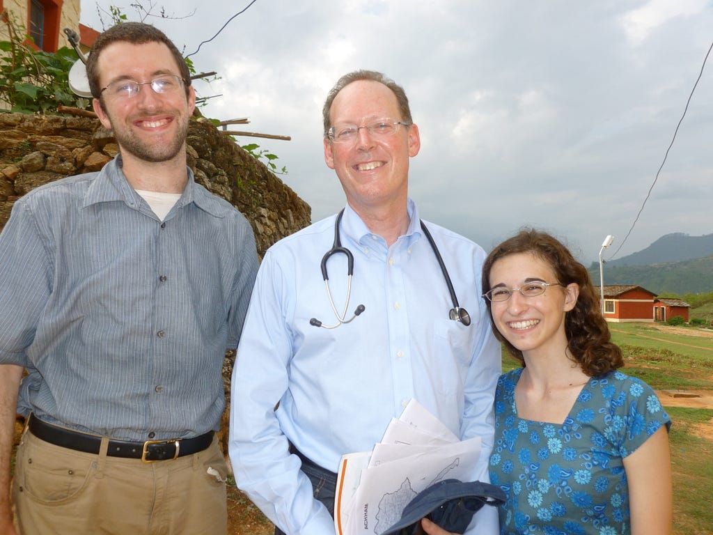 Paul Farmer stands between two GlobeMed students. He is wearing a long-sleeve, light blue button up shirt with a stethoscope slung around his neck. To the left is a student who is taller than Paul, has some scruffy facial hair, and is wearing a short-sleeve, gray button up. To the right is a student who is shorter than Paul and is wearing a blue patterned top with a square neckline. They are all smiling at the camera, with a rural landscape in the background.