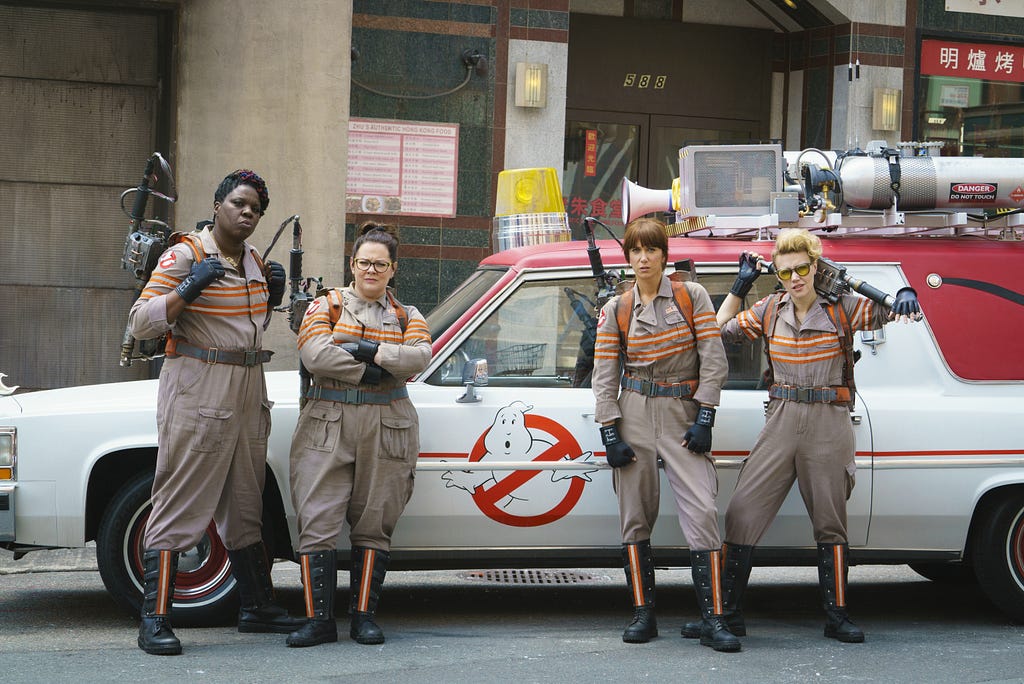 A promotional photo from Ghostbusters (2016) showing the four main cast-members stood beside their incarnation of the Ecto-1.