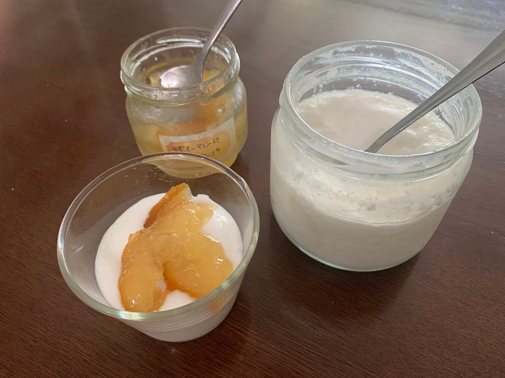 A glass jar of homemade yogurt with a spoon inside sits on a wooden table. Next to it is a small glass bowl containing yogurt topped with homemade yuzu marmalade. An open jar of the yuzu marmalade with a spoon is also present.